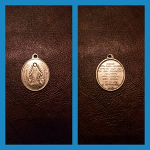 May include: A silver-colored religious pendant with a loop for hanging. One side features an image of a robed figure with a halo, surrounded by text. The other side has text in a circular pattern.