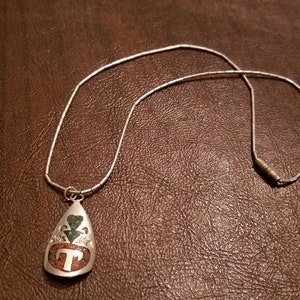 May include: A silver chain necklace with a teardrop-shaped pendant. The pendant is made of brown, green, and white stones and features a silver letter "T" in the center.