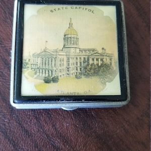 May include: A silver metal compact with a black border and a picture of the Georgia State Capitol building in Atlanta, Georgia. The text "STATE CAPITOL" is printed above the building and "ATLANTA, GA" is printed below the building.