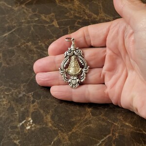 Op de afbeelding: Een zilveren en gouden hanger met een religieus beeld van de Maagd Maria. De hanger is versierd en heeft een gedetailleerd ontwerp.
