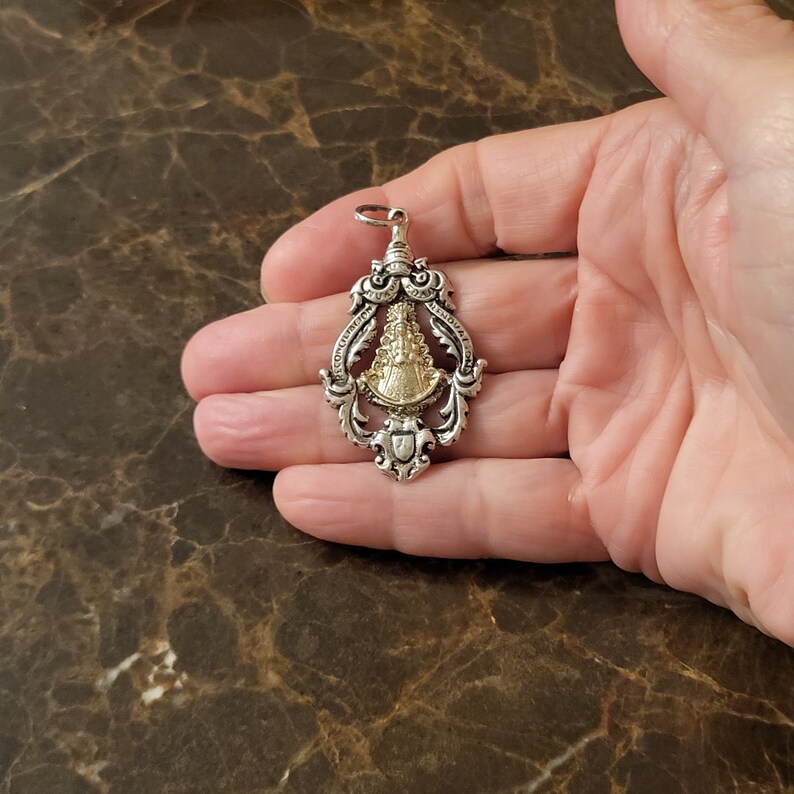 Op de afbeelding: Een zilveren en gouden hanger met een religieus beeld van de Maagd Maria. De hanger is versierd en heeft een gedetailleerd ontwerp.