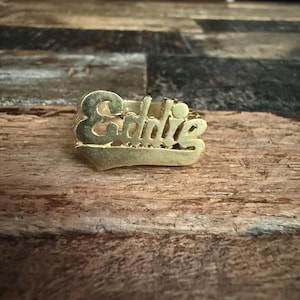 May include: A gold-colored metal ring featuring the name Eddie in a cursive script font. The name is centered on a horizontal bar design that forms the front face of the ring, with a polished metallic finish and visible lettering detail.
