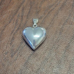 May include: A polished silver-toned heart pendant with a smooth, reflective surface and a small metal bail at the top for attachment. The heart shape is rounded and dimensional, featuring a clean, minimalist design without any additional engravings.
