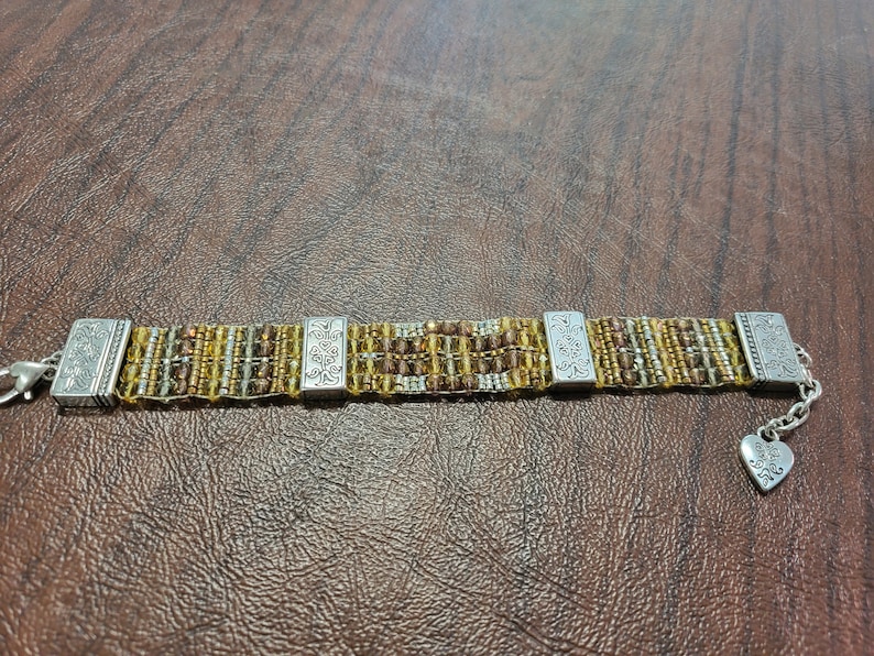 May include: A gold and silver beaded bracelet with silver end caps and a heart charm. The bracelet has a floral design on the end caps.