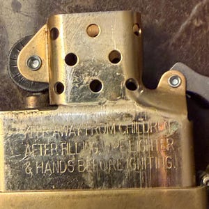 K&ouml;nnte beinhalten: Ein messingfarbener Feuerzeugeinsatz aus Metall mit perforiertem Windschutz und Z&uuml;ndrad. Die Oberfl&auml;che ist mit folgendem Text graviert: KEEP AWAY FROM CHILDREN. AFTER FILLING, WIPE LIGHTER & HANDS BEFORE IGNITING.