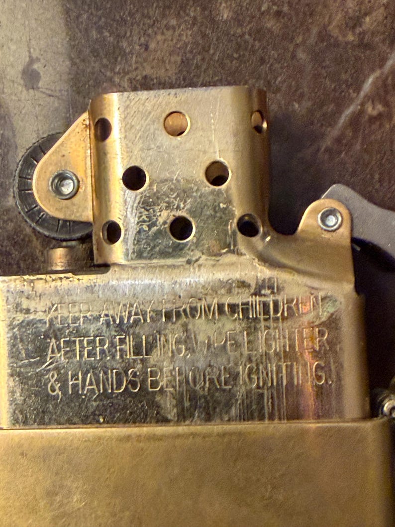 K&ouml;nnte beinhalten: Ein messingfarbener Feuerzeugeinsatz aus Metall mit perforiertem Windschutz und Z&uuml;ndrad. Die Oberfl&auml;che ist mit folgendem Text graviert: KEEP AWAY FROM CHILDREN. AFTER FILLING, WIPE LIGHTER & HANDS BEFORE IGNITING.