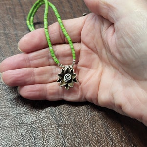 K&ouml;nnte beinhalten: Eine Halskette aus gr&uuml;nen Edelsteinperlen mit einem silbernen Blumenanh&auml;nger. Die Blume hat ein spiralf&ouml;rmiges Design in der Mitte.