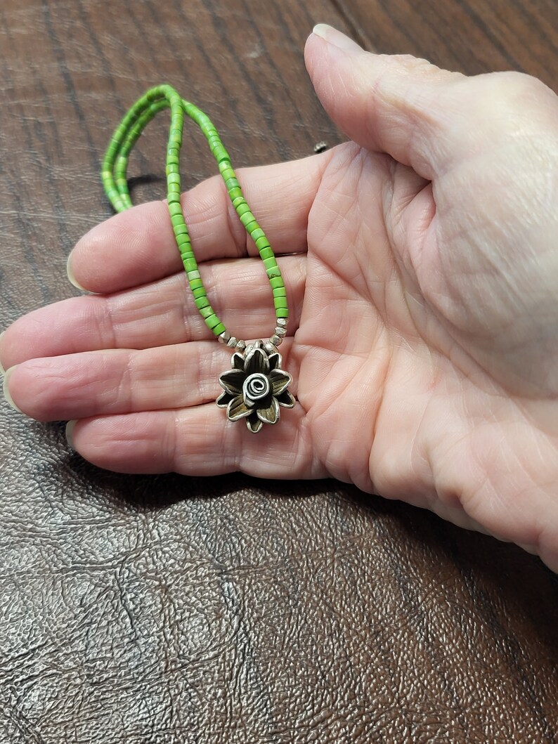 K&ouml;nnte beinhalten: Eine Halskette aus gr&uuml;nen Edelsteinperlen mit einem silbernen Blumenanh&auml;nger. Die Blume hat ein spiralf&ouml;rmiges Design in der Mitte.