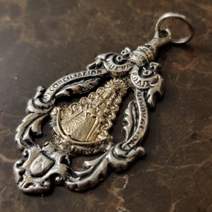 Op de afbeelding: Zilveren hanger met een goudkleurig beeld van de Maagd Maria. De hanger is versierd met sierlijke krulwerk en de woorden "NUEVA EVANGELIZACION" en "RECONCILIACION".