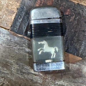May include: Vintage silver-toned lighter with a black band and a clear panel featuring a white horse silhouette. The lighter has a rectangular shape and a flip-top design. The lighter is on a wooden surface.