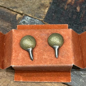 May include: A pair of golf ball cufflinks with a textured gold-tone ball design and silver-tone tee post. The cufflinks are presented in an open, orange velvet box, set against a rustic wooden surface.