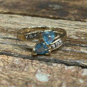 May include: A gold ring featuring two light blue, triangular gemstones. The ring has a band with small, clear accent stones. The ring is set against a textured, brown wooden surface.