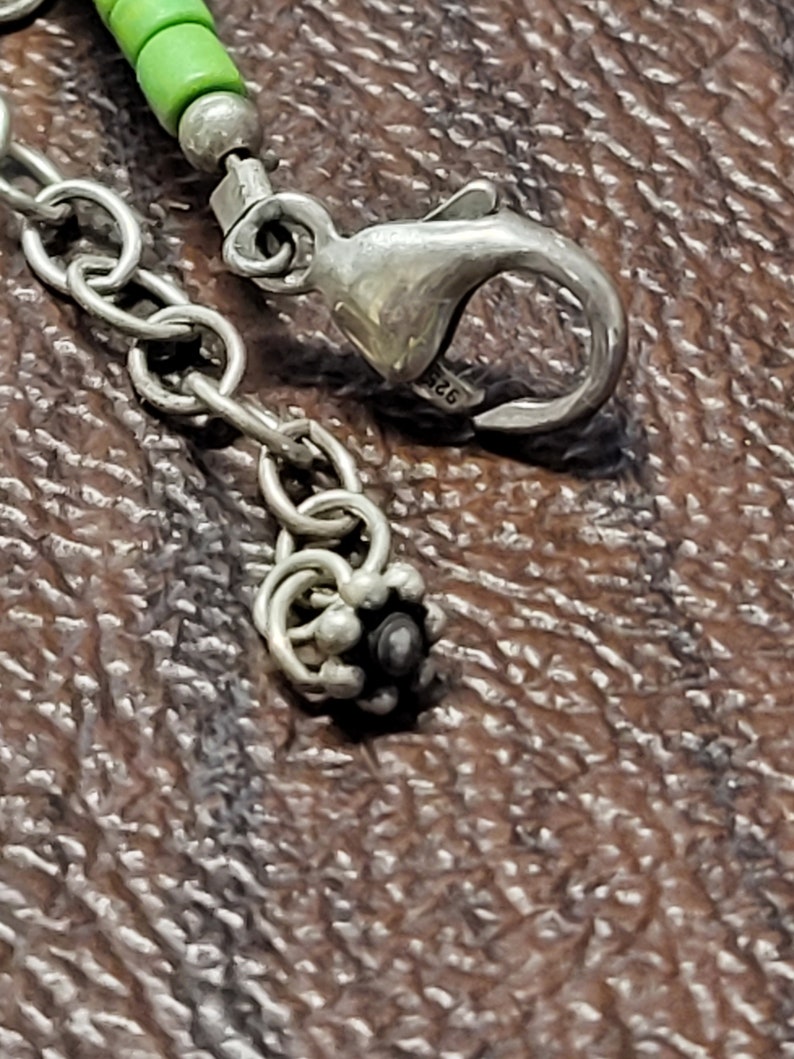 K&ouml;nnte beinhalten: Nahaufnahme eines silbernen Kettenarmbands mit einem Karabinerverschluss und einer dekorativen Perle. Die Perle ist silberfarben mit einem schwarzen Zentrum.