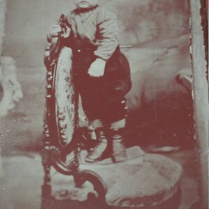 May include: A black and white vintage photograph of a young child sitting on a wooden chair. The child is wearing a sweater and pants. The photo is faded and has a brown tint.