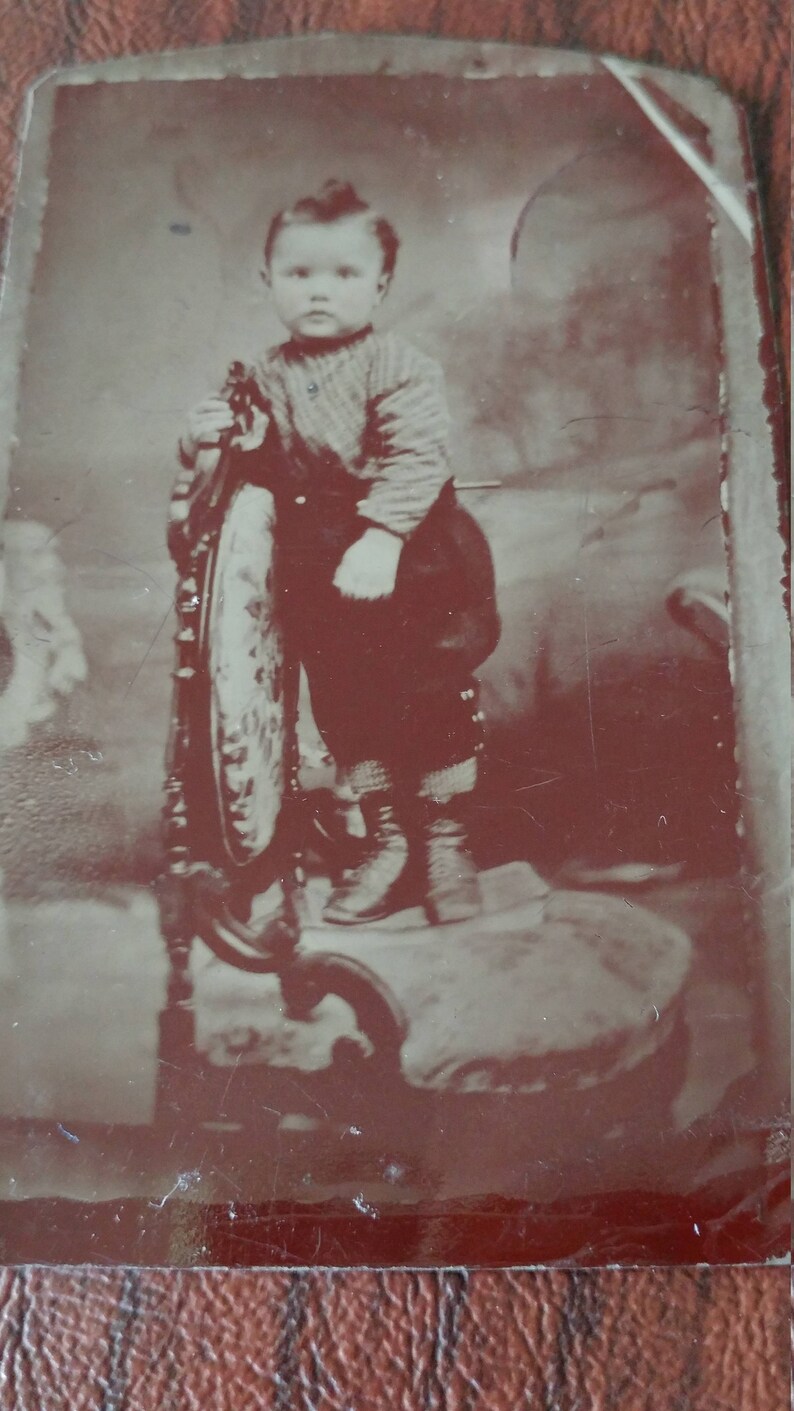 May include: A black and white vintage photograph of a young child sitting on a wooden chair. The child is wearing a sweater and pants. The photo is faded and has a brown tint.