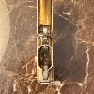 K&ouml;nnte beinhalten: Ein goldfarbenes Metallfeuerzeug mit Klappdeckel in ge&ouml;ffneter Position. Das Feuerzeug hat einen rechteckigen K&ouml;rper mit einem Klappdeckel, einen zentralen Kamin mit sichtbarem wei&szlig;em Docht und eine metallische Innenoberfl&auml;che.
