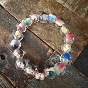 May include: A circular bracelet composed of clear glass beads, each featuring a hand-painted miniature face with distinct expressions and colorful accents in red, blue, green, and purple, strung together to form a continuous decorative loop.