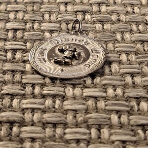 May include: A circular silver-toned metal pendant featuring a raised relief of Mickey Mouse in the center. The text Walt Disney World is embossed along the outer edge of the circular frame, which has a textured border and a small metal loop at the top.