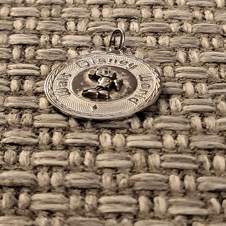 May include: A circular silver-toned metal pendant featuring a raised relief of Mickey Mouse in the center. The text Walt Disney World is embossed along the outer edge of the circular frame, which has a textured border and a small metal loop at the top.