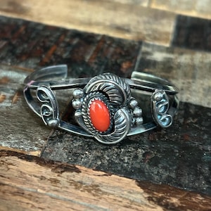 May include: A silver cuff bracelet with an oval, bright red stone at the center. The stone is surrounded by ornate silver detailing, including leaf and bead designs. The bracelet is displayed on a wooden surface.