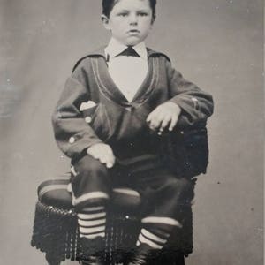 May include: A black and white portrait of a young boy wearing a sailor suit with striped socks and black shoes. He is sitting on a chair with a fringed cushion.