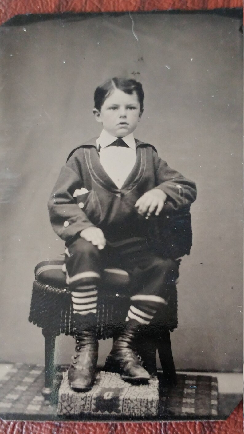 May include: A black and white portrait of a young boy wearing a sailor suit with striped socks and black shoes. He is sitting on a chair with a fringed cushion.