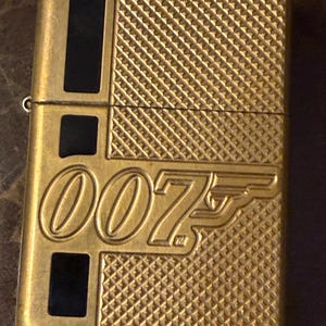 K&ouml;nnte beinhalten: Ein goldfarbenes Feuerzeug aus Metall mit dem gepr&auml;gten 007-Logo auf der Vorderseite. Die Oberfl&auml;che weist auf der rechten Seite ein strukturiertes Kreuzschraffurmuster und auf der linken Seite drei vertikale schwarze Rechteckeinlagen auf.