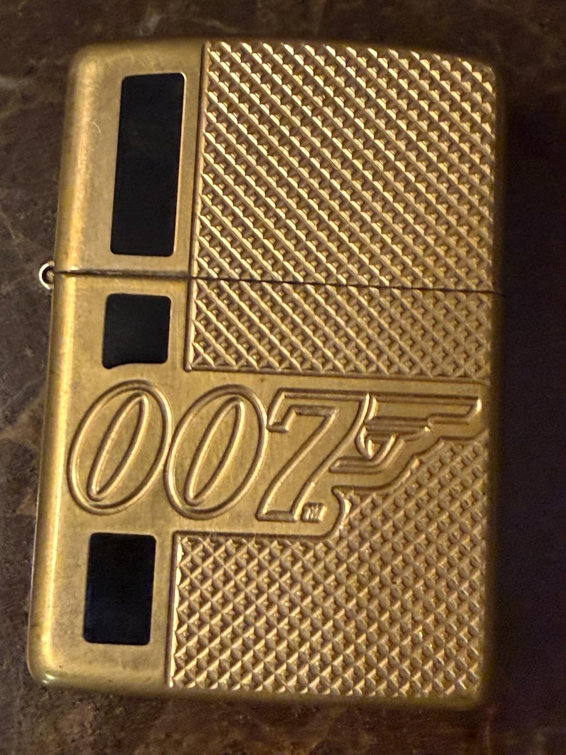 K&ouml;nnte beinhalten: Ein goldfarbenes Feuerzeug aus Metall mit dem gepr&auml;gten 007-Logo auf der Vorderseite. Die Oberfl&auml;che weist auf der rechten Seite ein strukturiertes Kreuzschraffurmuster und auf der linken Seite drei vertikale schwarze Rechteckeinlagen auf.