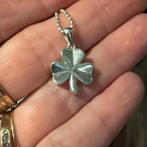 May include: A silver-toned shamrock pendant suspended from a delicate ball chain. The three-leaf clover charm features a polished, reflective surface with a smooth finish, showcasing a classic botanical shape attached to a small metal bail.