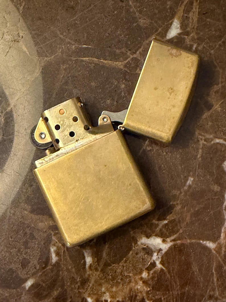 K&ouml;nnte beinhalten: Ein rechteckiges, messingfarbenes Metallfeuerzeug mit offenem Klappdeckel. Das Feuerzeug verf&uuml;gt &uuml;ber einen metallischen Kamin mit kreisf&ouml;rmigen Bel&uuml;ftungsl&ouml;chern und einen kleinen Reibradmechanismus an der Seite des inneren Einsatzes.
