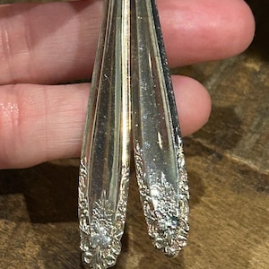 May include: Silver-toned, vintage-style earrings. Each earring is crafted from a repurposed spoon, featuring a floral design at the base. The earrings have a teardrop shape and are suspended from a small loop.