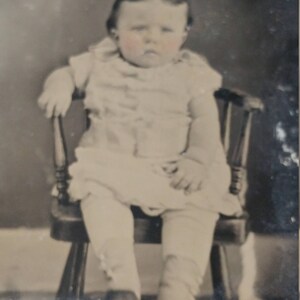 May include: A black and white photo of a young child sitting in a wooden chair. The child is wearing a white dress and has a serious expression on their face.
