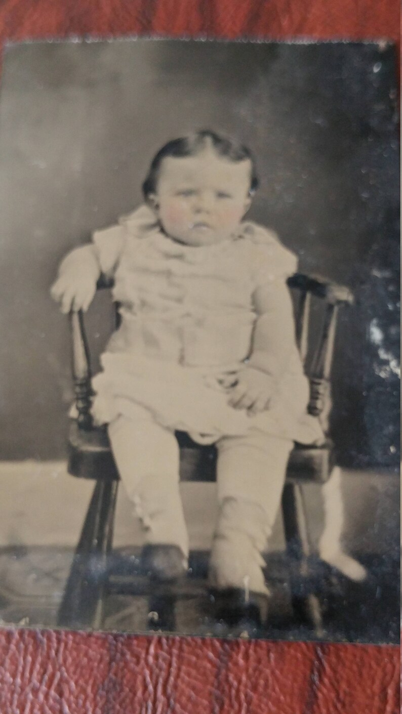 May include: A black and white photo of a young child sitting in a wooden chair. The child is wearing a white dress and has a serious expression on their face.