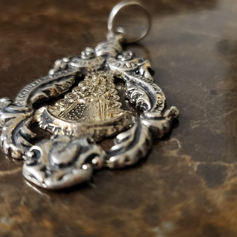 Op de afbeelding: Zilveren hanger met een gouden afbeelding van een religieuze figuur. De hanger is versierd en heeft ingewikkelde details.
