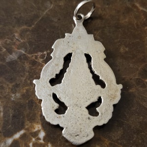 Op de afbeelding: Zilveren hanger met een gedetailleerd, sierlijk ontwerp. De hanger heeft een lus aan de bovenkant om aan een ketting te bevestigen.