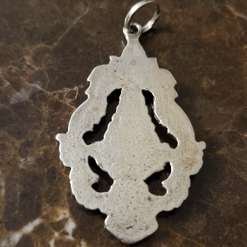 Op de afbeelding: Zilveren hanger met een gedetailleerd, sierlijk ontwerp. De hanger heeft een lus aan de bovenkant om aan een ketting te bevestigen.