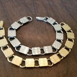 May include: A gold and silver-toned bracelet and necklace set. The jewelry features a repeating pattern of square links with a textured, diamond-like design. The necklace is connected with a clasp, and the bracelet is next to it.