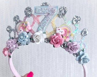 Puede incluir: Una diadema rosa con una corona blanca decorada con purpurina plateada, pedrería, flores pastel y un amuleto de unicornio arcoíris. La corona presenta un número 7 brillante.