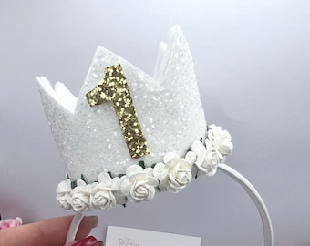 Op de afbeelding: Een witte glitter kroon hoofdband met een gouden glitter nummer 1. De kroon is versierd met kleine witte bloemen en zit op een witte hoofdband. Een visitekaartje met de tekst "Hallebellasbling.com" is zichtbaar.