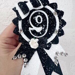 May include: Black and white glitter rosette with a large number nine in the center. The rosette has a white and black bow with rhinestones and a white and black flower.