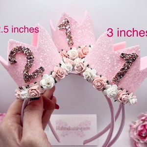 May include: Two pink glitter crowns with white and pink flowers, one with the number "1" and the other with the number "2", on a pink headband. The crowns are 2.5 inches and 3 inches tall.