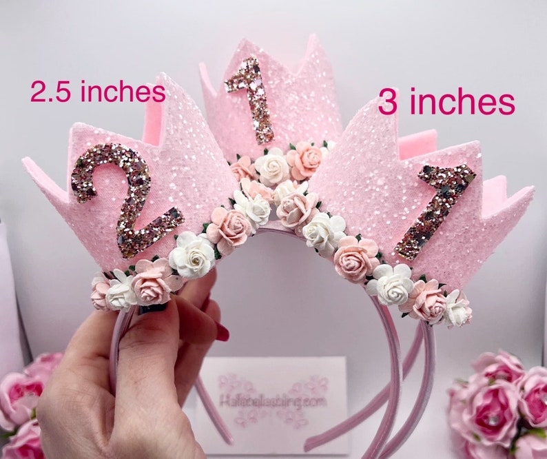 May include: Two pink glitter crowns with white and pink flowers, one with the number "1" and the other with the number "2", on a pink headband. The crowns are 2.5 inches and 3 inches tall.