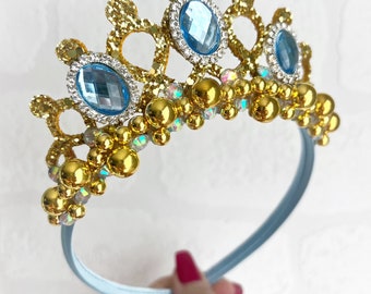 May include: A gold crown headband with blue gem accents and gold beads. The headband is light blue.