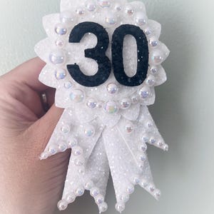 May include: A white, glittery rosette-style badge with the number 30 in black. The badge is adorned with iridescent pearls and ribbon tails, perfect for a milestone birthday celebration.