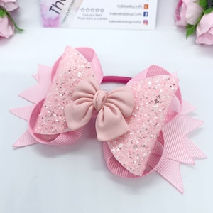 Peut inclure: Noeud pour cheveux en paillettes rose avec un grand noeud en satin rose au centre. Le noeud est attaché à un élastique à cheveux rose.