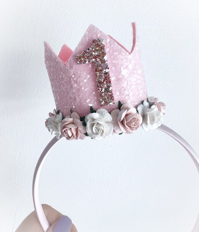 May include: Pink felt crown headband with a glittery number one and a row of white and pink flowers around the base.