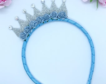 May include: A light blue headband with a silver glitter crown and blue fabric flowers.