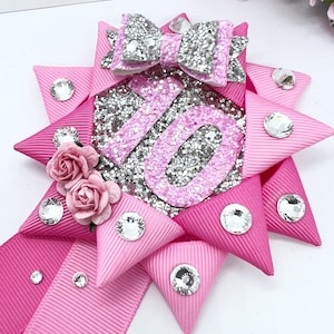 May include: Pink and silver glitter bow with the number 16 in the center. The bow is decorated with rhinestones and small pink flowers.