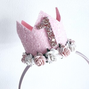 May include: Pink felt crown with a glittery number one and a floral headband. The crown is decorated with white and pink felt flowers.
