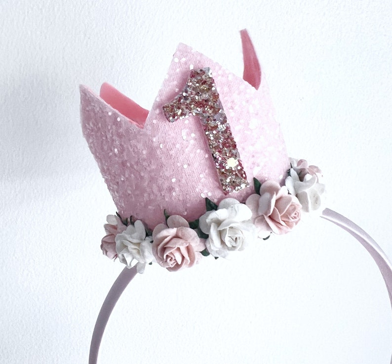 May include: Pink felt crown with a glittery number one and a floral headband. The crown is decorated with white and pink felt flowers.